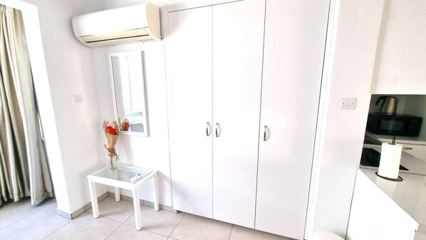 A white wardrobe with two doors, a small table with a vase of flowers, and a kitchen area with a microwave and a kettle.