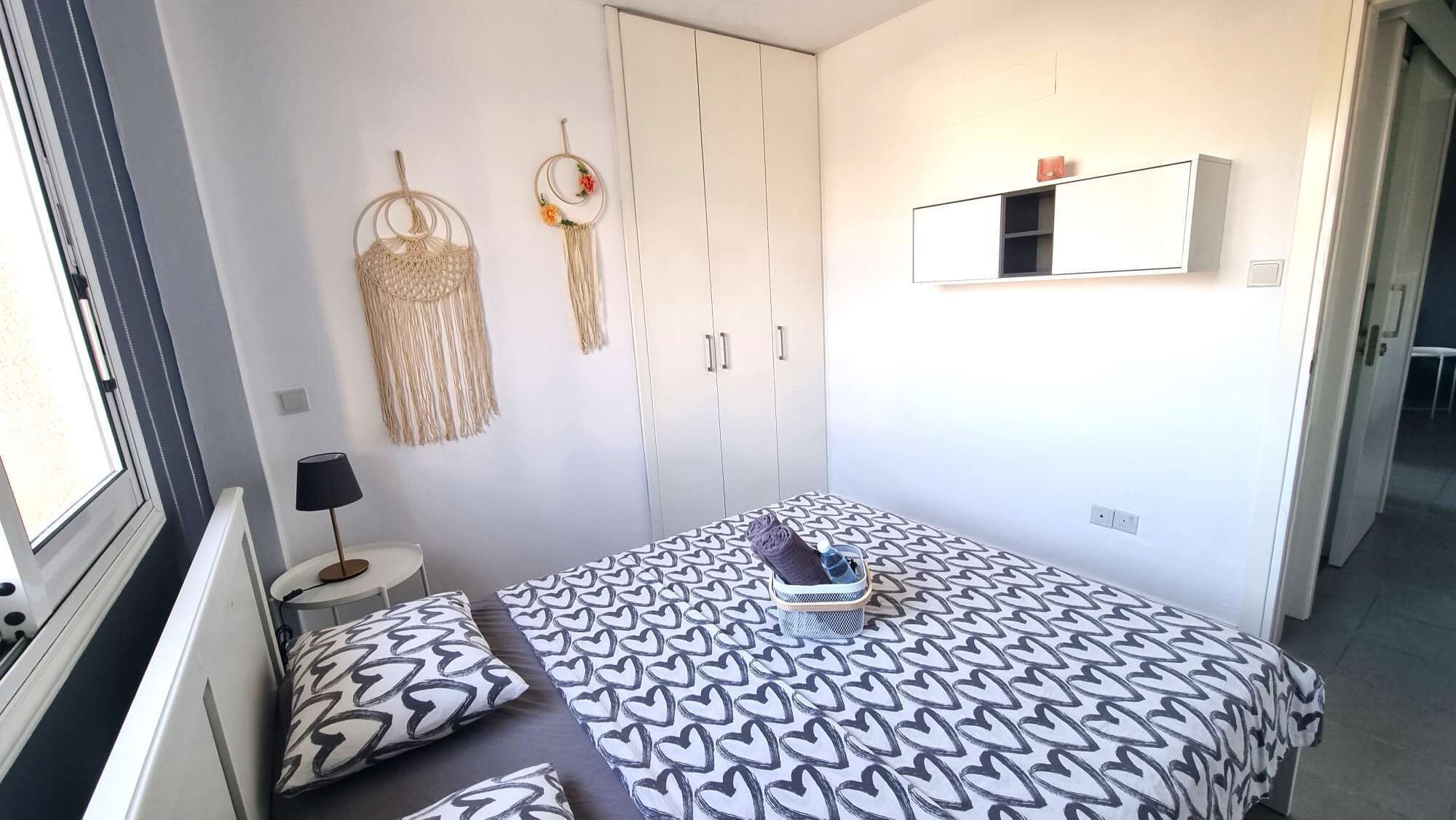 Ein Schlafzimmer mit einem Bett, einem Kleiderschrank, einem kleinen Tisch mit Lampe sowie Wandbehängen aus Makramee.