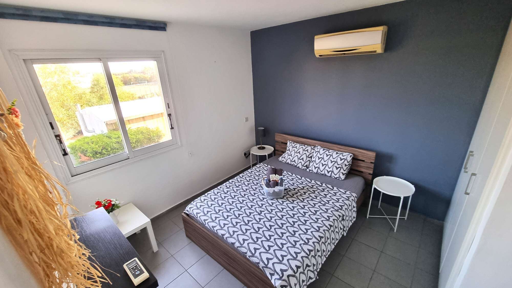 Une chambre à coucher comprenant un lit, des tables de chevet, une fenêtre et un appareil de climatisation.