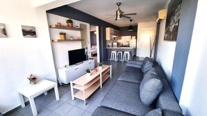 Een moderne woonkamer met een grijze bank, een televisie, een witte salontafel en een klein witte tafeltje met een vaas. Op de achtergrond is een keuken te zien met een bar en krukken.