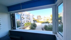 Een kamer met een groot raam met uitzicht op een woonwijk, met een blauw luifel en een zicht op meer verdiepingen tellende gebouwen.