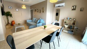 A modern living room with a dining table, sofa, and chairs. There are two pendant lights, a TV, and a bookshelf. - 2Br Apt - 5mins walk to the Beach + Netflix access (Larnaca)