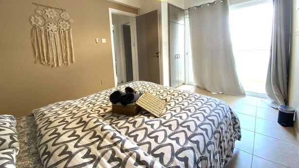 A bedroom with a patterned bedspread, a hanging light fixture, a dreamcatcher on the wall, and a view of the outside through a window with curtains. - 2Br Apt - 5mins walk to the Beach + Netflix access (Larnaca)