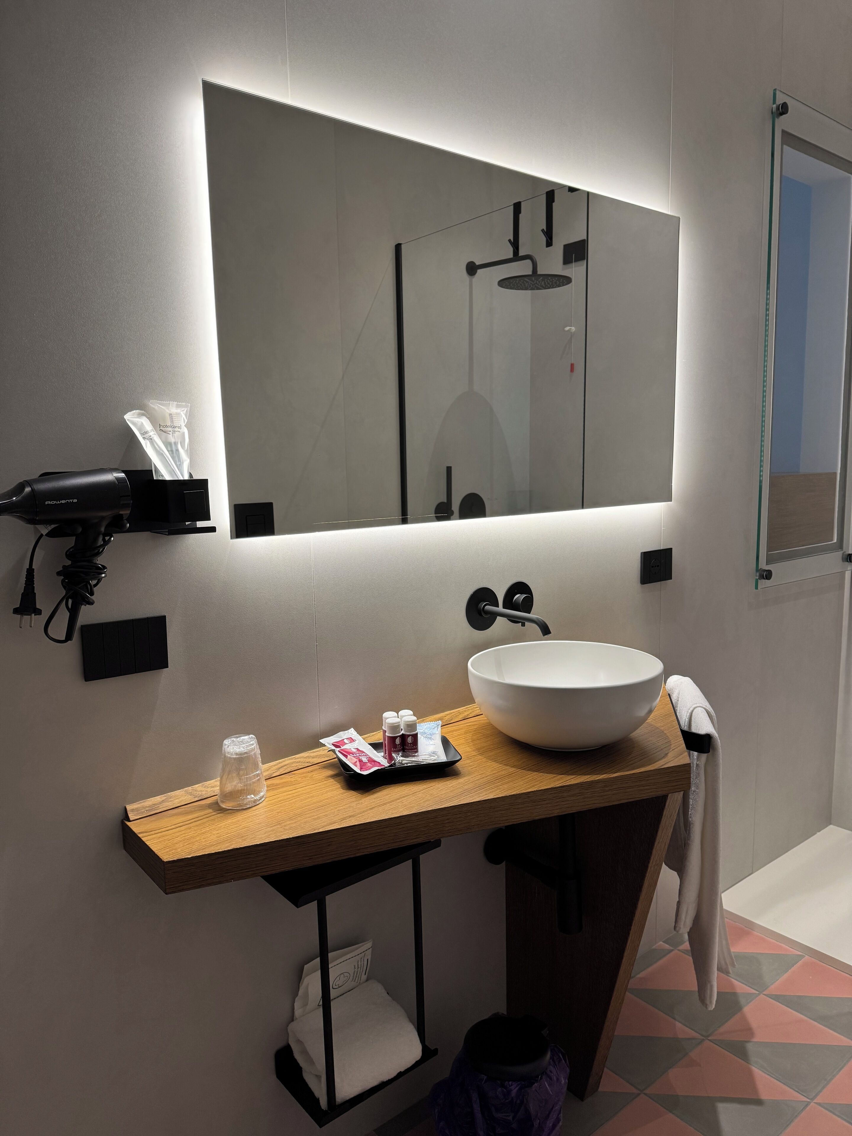 A modern bathroom with a large mirror, a wooden vanity, a white sink, and a towel hanging on the wall.