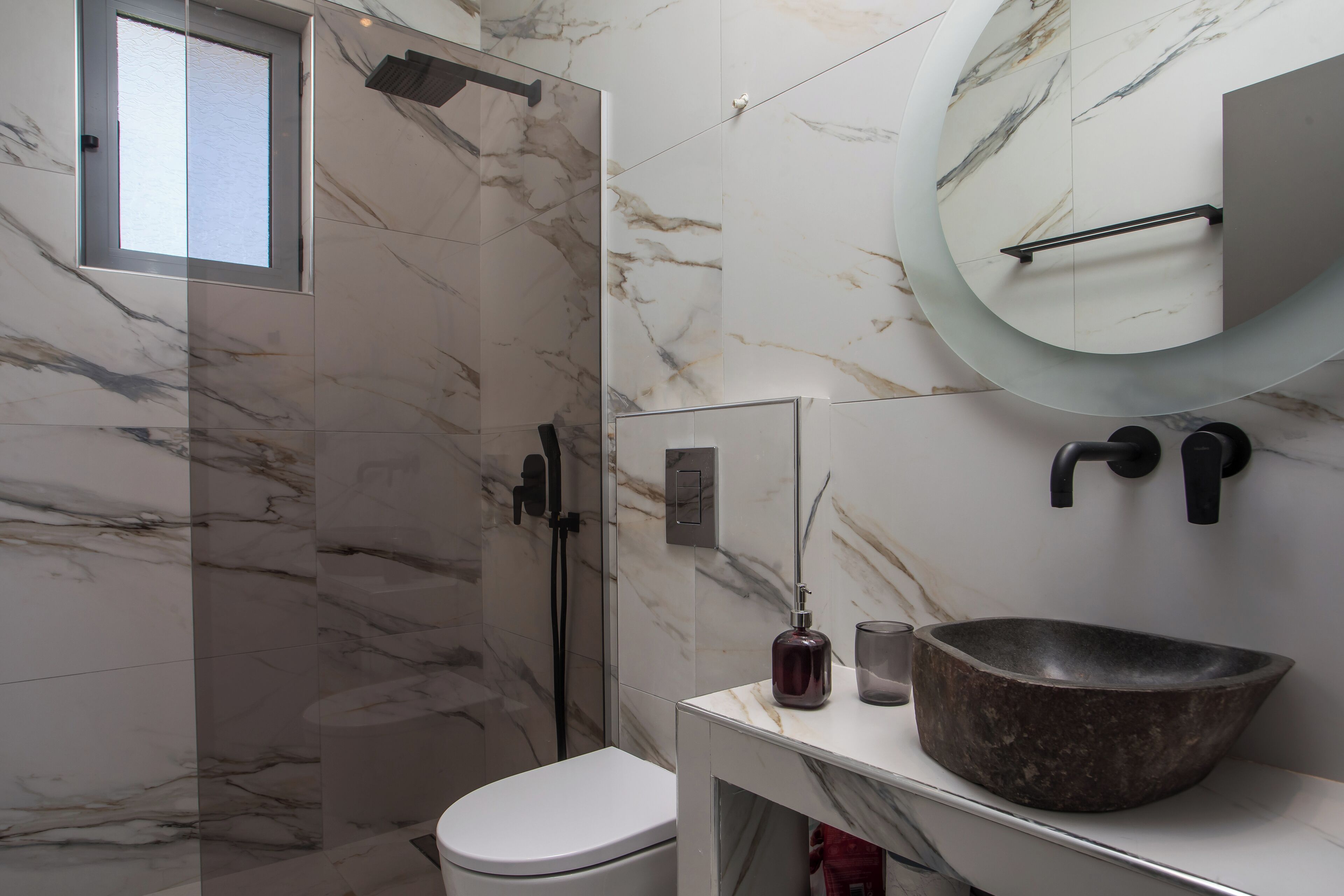 Une salle de bain avec un mur en marbre, des toilettes blanches, un grand lavabo sur pied et une douche à parois de verre.