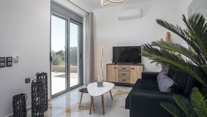 A modern living room with a large sliding glass door, a flat-screen TV, a dark sofa, and a small white table. - Marinas villas (Rhodes)