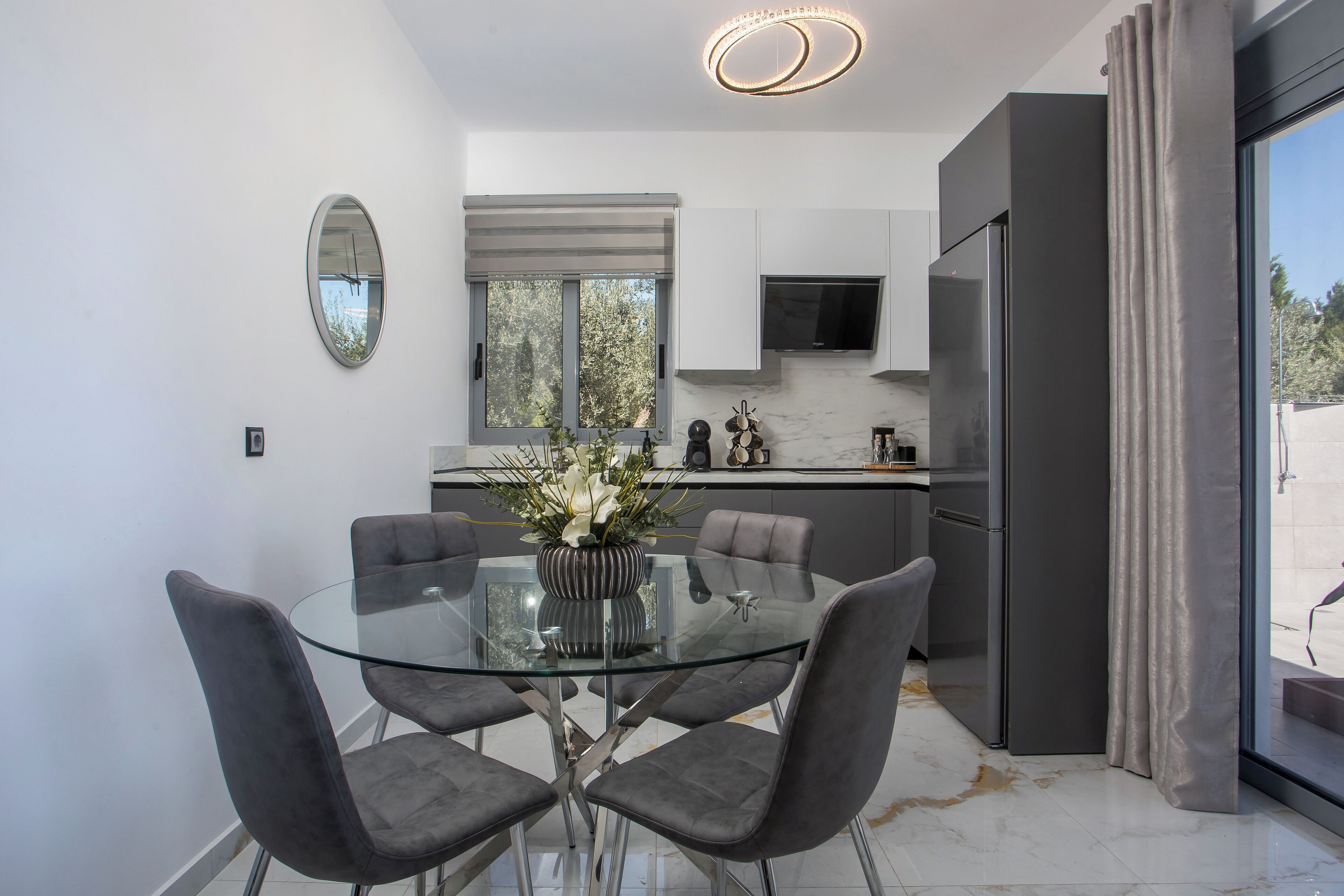 Una cocina moderna con una mesa de comedor de cristal, sillas grises y un ventanal con vistas a árboles.