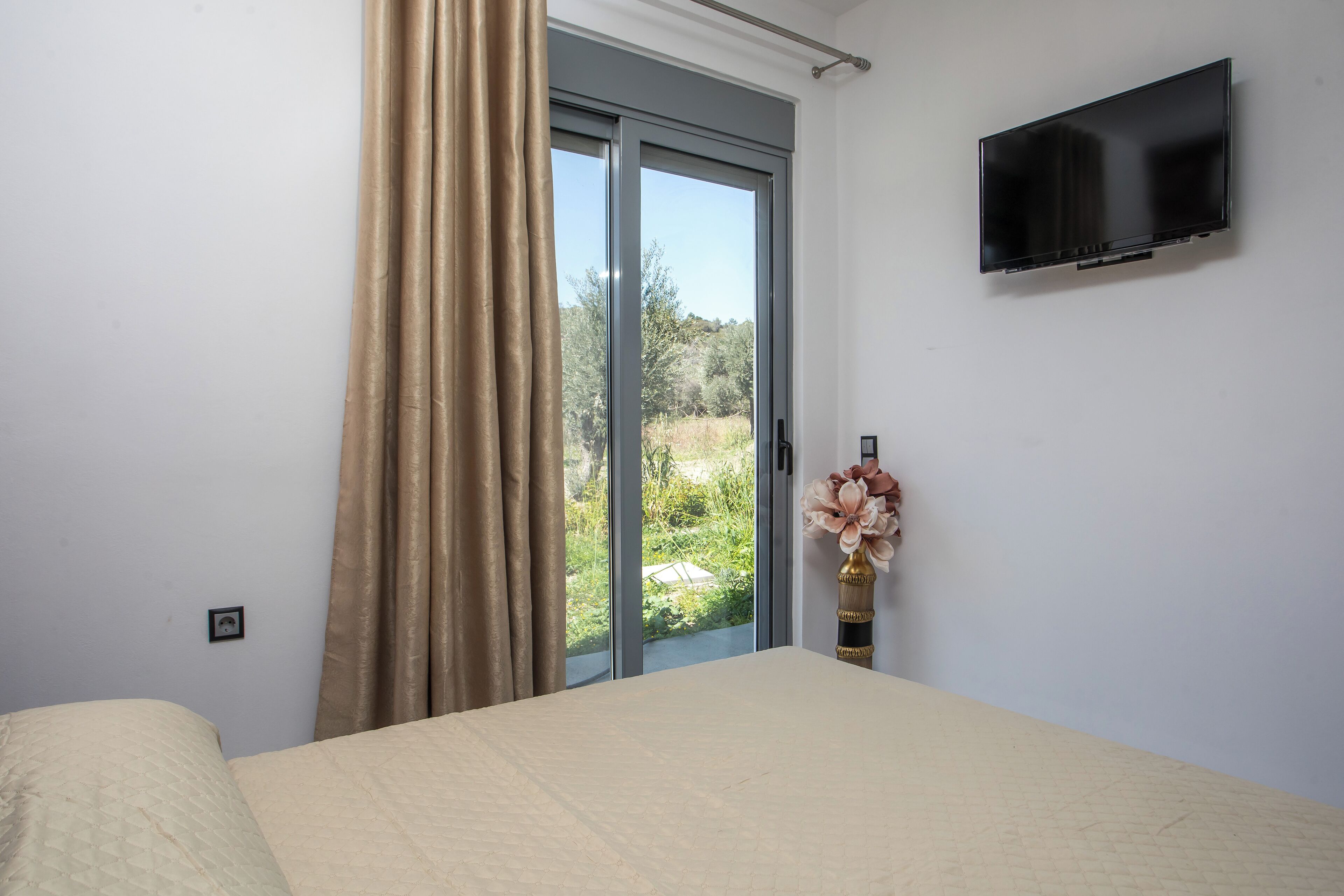 A bedroom with a bed, a TV mounted on the wall, a window with curtains, and a vase with flowers on the windowsill.