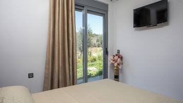 A bedroom with a bed, a TV mounted on the wall, a window with curtains, and a vase with flowers on the windowsill.
