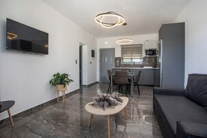 A modern living room with a flat-screen TV, a dining area with a table and chairs, and a kitchen with a microwave and refrigerator.