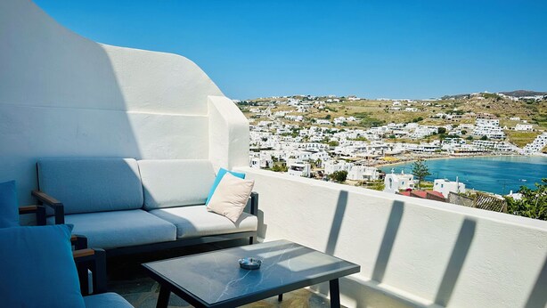 A balcony with a sofa and table, overlooking a coastal town with white buildings.