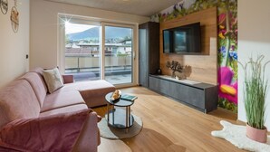 Een moderne woonkamer met een bank, een salontafel, een aan de muur bevestigde televisie en een balkon met uitzicht op gebouwen en bergen.