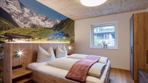 Un dormitorio con una cama de madera, un mural de montañas en la pared, una ventana con vista a un edificio y una lámpara en el techo.
