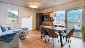 Una sala de estar moderna con techo de madera, un comedor con mesa y sillas, un sofá y vistas a un pueblo a través de ventanas amplias.