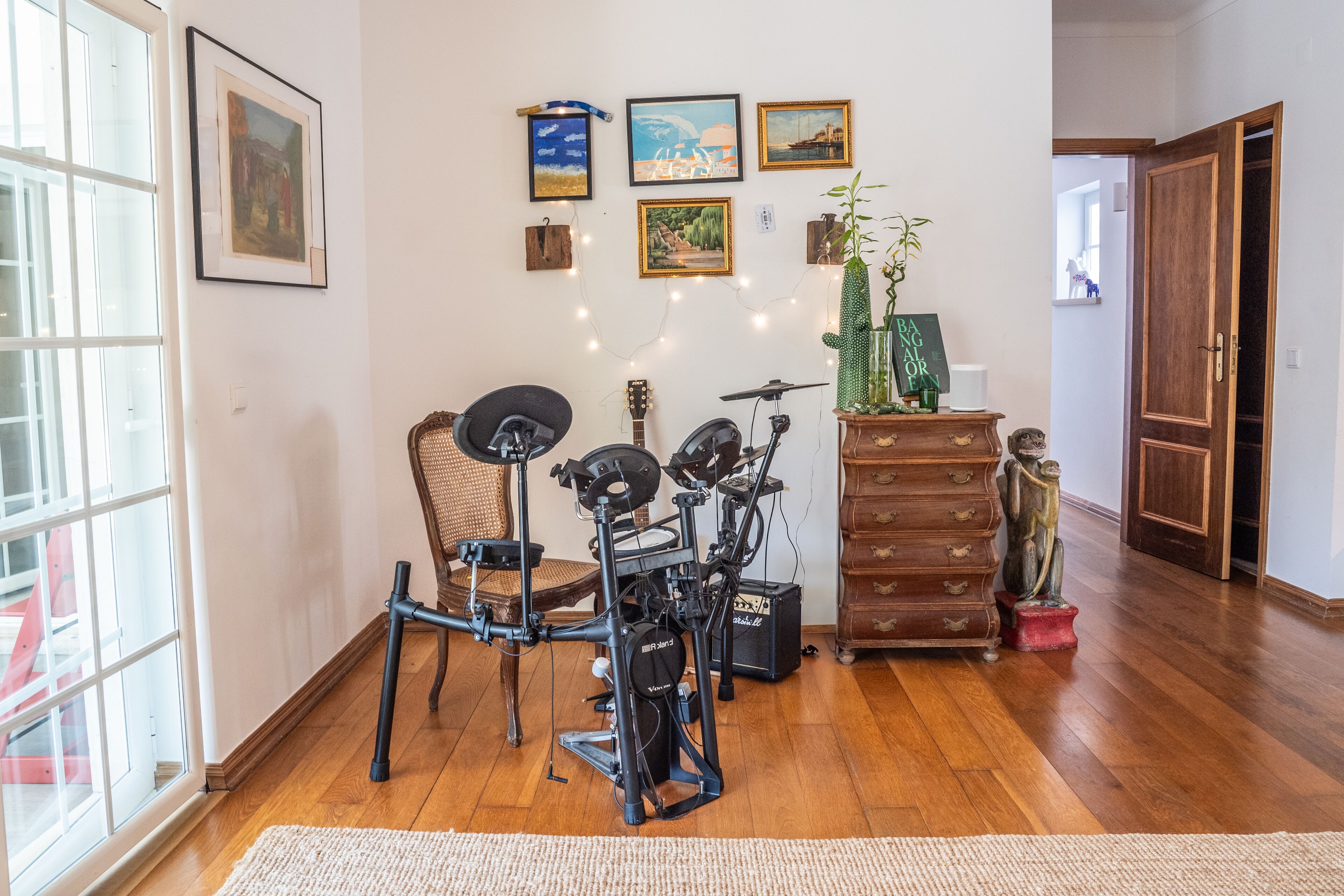 A room with a wooden floor, a large window, a chair, a guitar, a drum set, a dresser, and several framed pictures on the wall.
