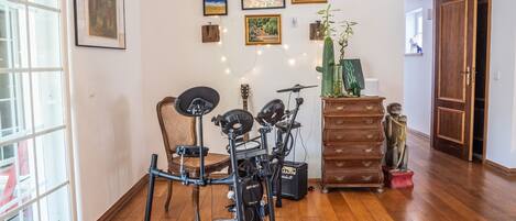 A room with a wooden floor, a large window, a chair, a guitar, a drum set, a dresser, and several framed pictures on the wall.