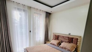 Une chambre à coucher avec un grand lit, une table de chevet, un luminaire au plafond et une fenêtre avec des rideaux.