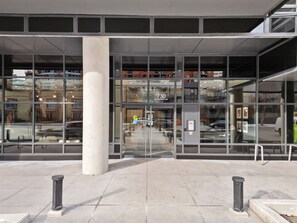 Une entrée moderne, caractérisée par de grandes fenêtres en verre, une enseigne indiquant l’adresse bien visible et un pilier en béton.