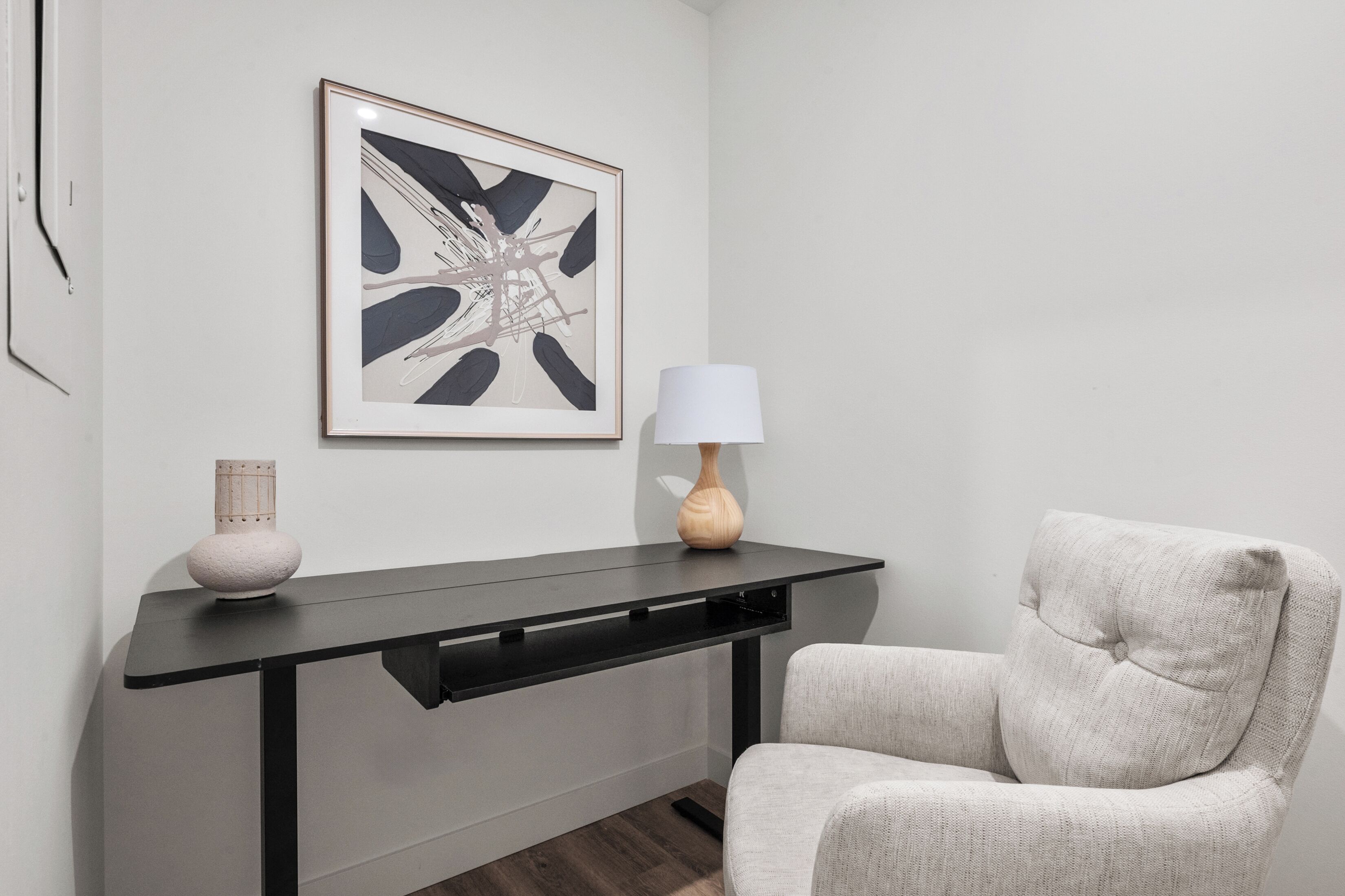 Una habitación moderna con un escritorio negro, una silla, una obra de arte abstracta enmarcada y un jarrón.