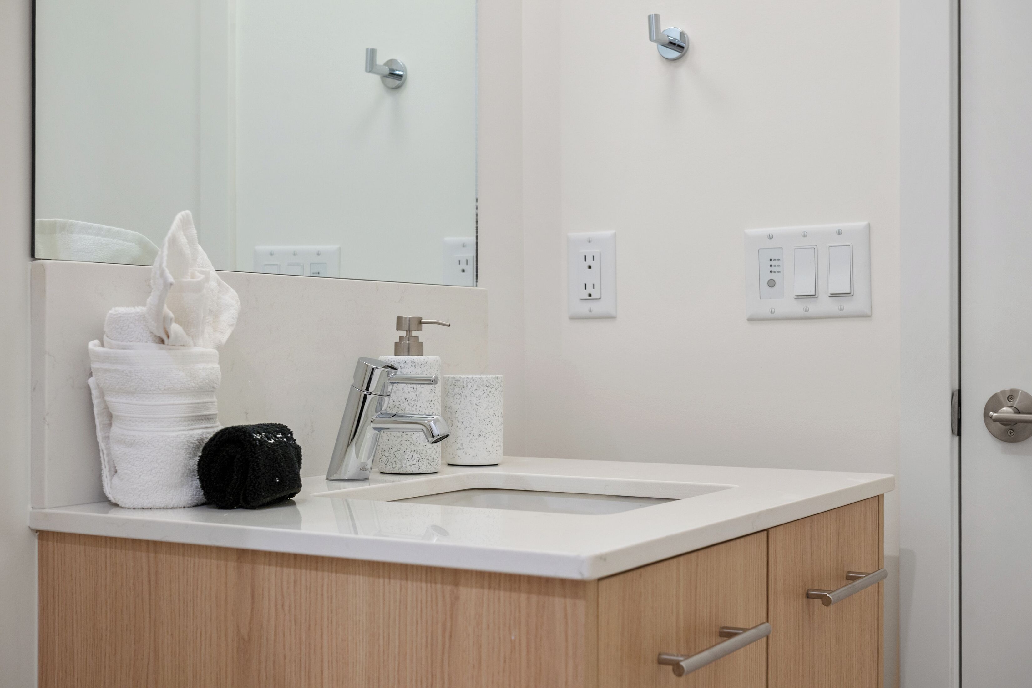 Un baño con un lavabo blanco, un espejo, una toalla, un bolso negro y un dispensador de jabón.