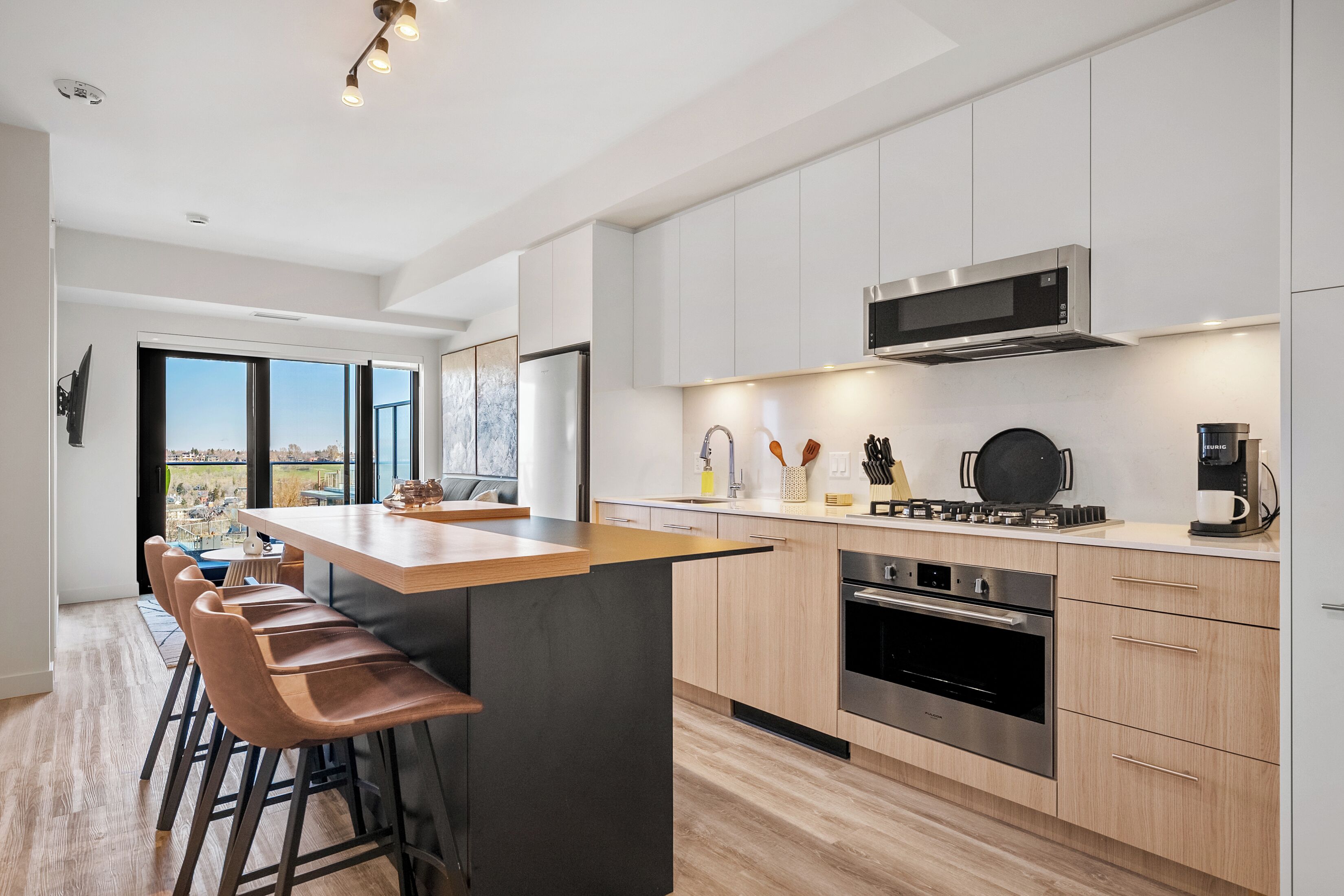 A modern kitchen with a central island, bar stools, stainless steel appliances, and a view through large windows.
