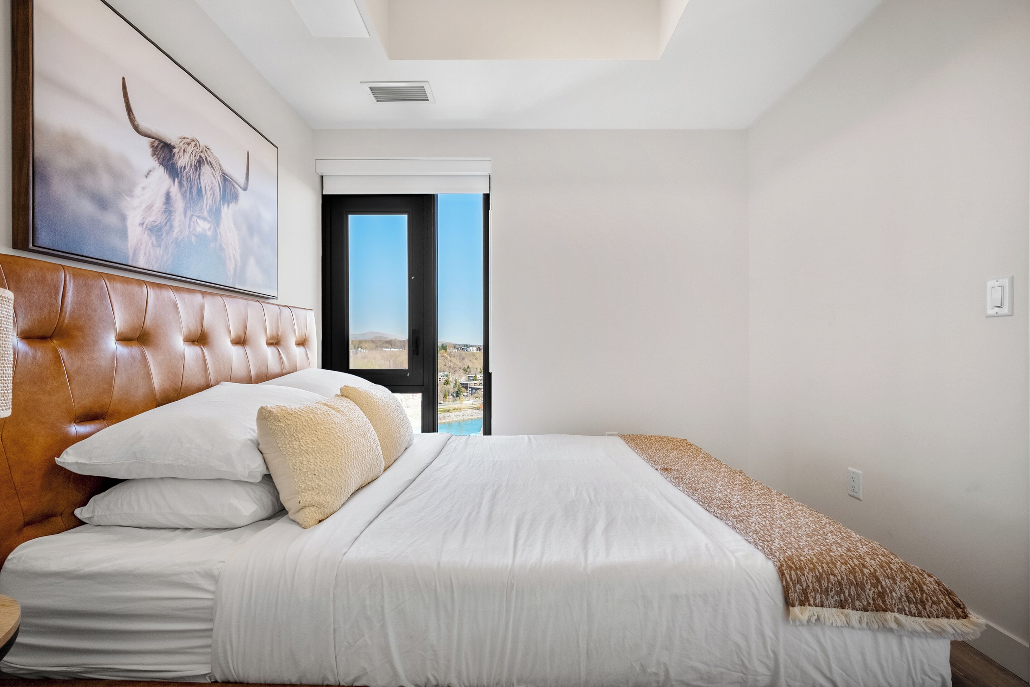 A bedroom with a large bed, a headboard, a framed picture, and a view of the outdoors.