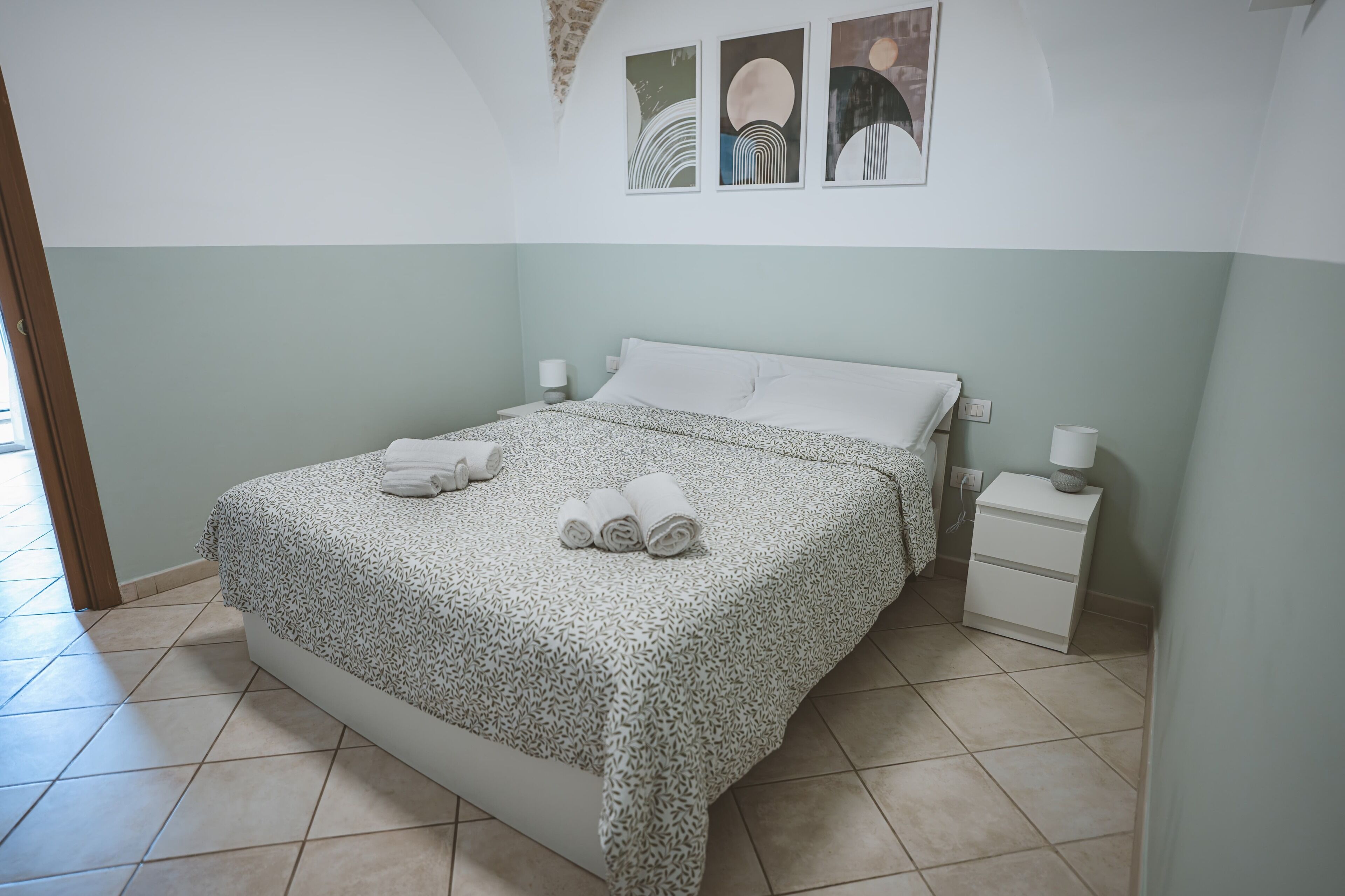 Una cama bien tendida con una colcha estampada y toallas enrolladas, una mesita de noche con lámpara, y una pared con arte abstracto.