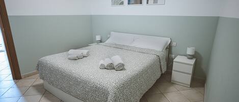 Una cama bien tendida con una colcha estampada y toallas enrolladas, una mesita de noche con lámpara, y una pared con arte abstracto.