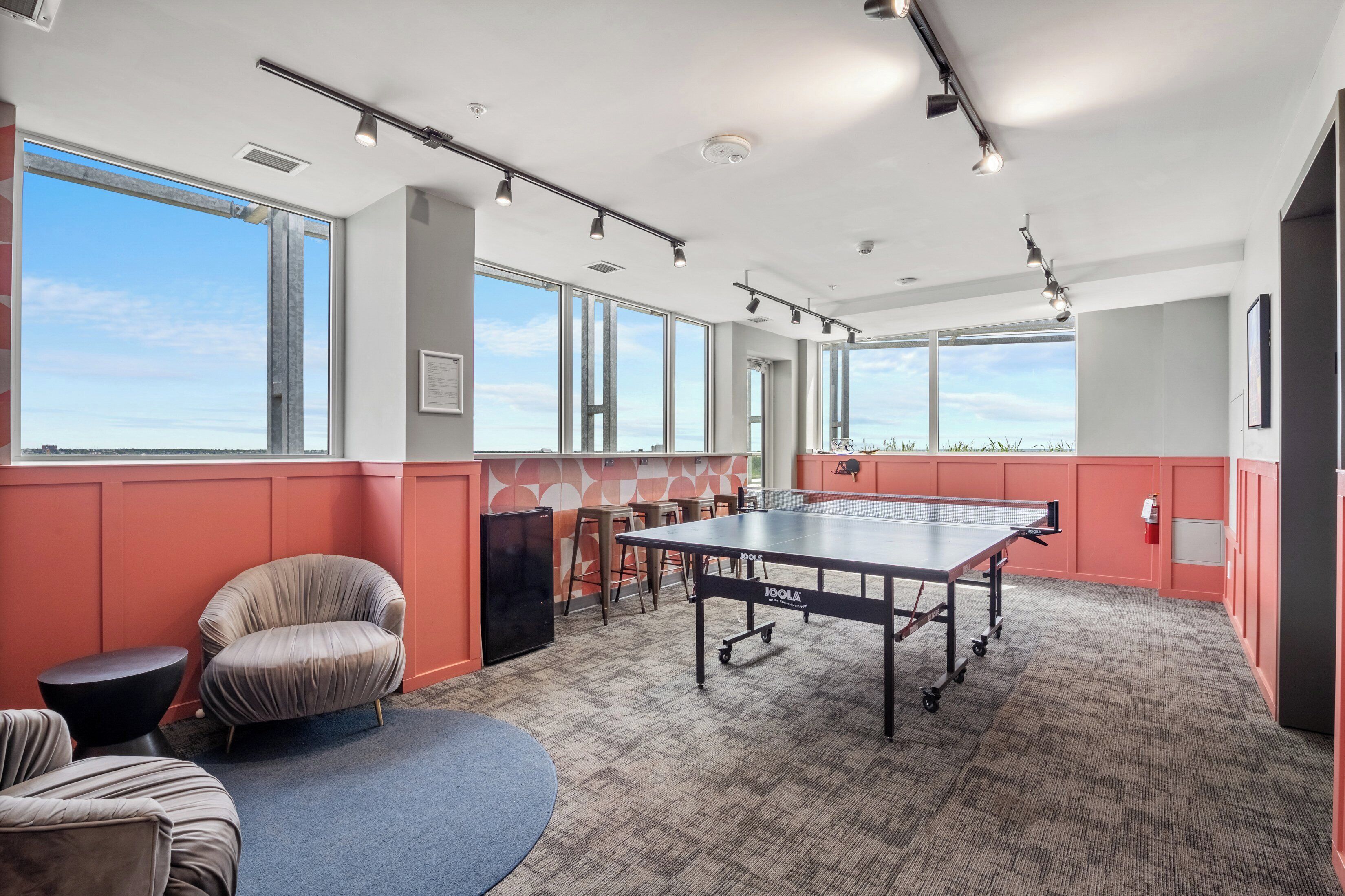Une pièce spacieuse équipée d’une table de ping-pong, de sièges confortables, de grandes fenêtres et d’un sol recouvert de moquette.