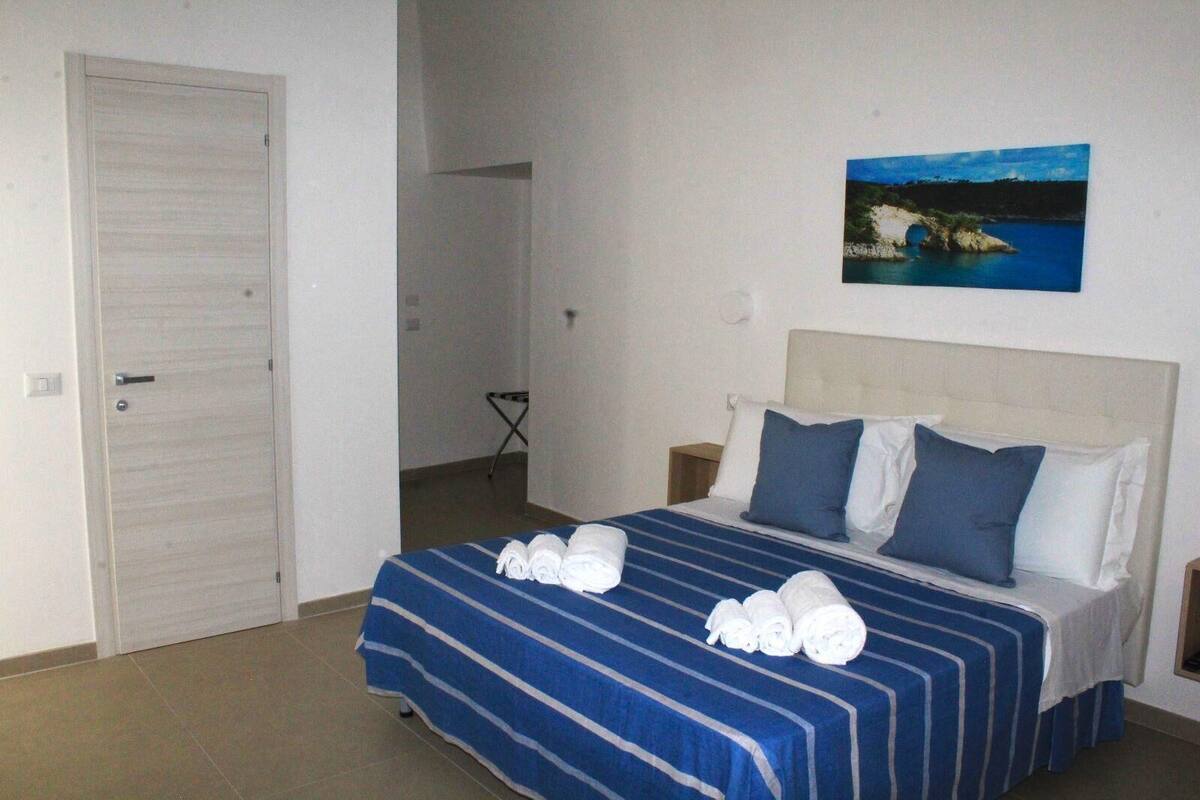 A hotel room with a bed, a painting of a coastal scene, and a door.
