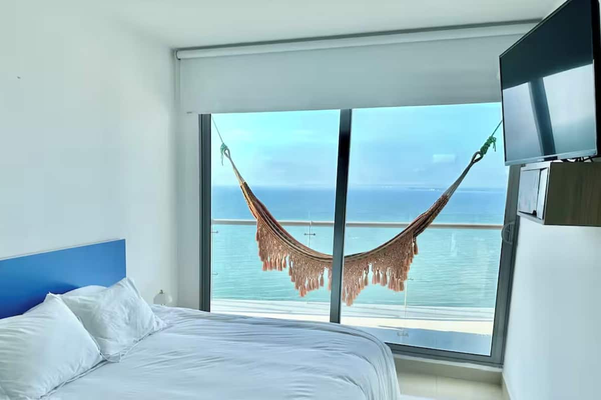 Quarto de hotel com uma cama, televisão e vista para o mar através de uma grande janela.