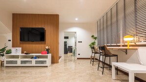 A modern living room with a flat-screen TV mounted on a wooden panel, a white entertainment unit, a dining area with a table and chairs, and a bench.