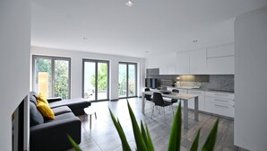 Une cuisine moderne avec des meubles blancs, un coin repas comprenant une table et des chaises, et un salon avec un canapé et une plante.