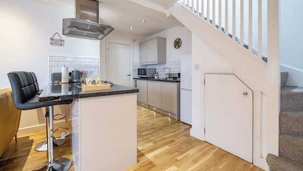 A modern kitchen with a central island, a dining area with a bar stool, and a staircase leading to an upper level. - Seaside Cottage Escape in Paignton - Sleeps 4 (Paignton)