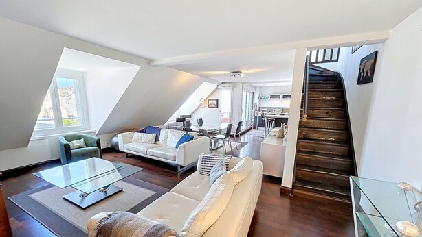 Un loft moderno con sala de estar que incluye un sofá blanco, una mesita de centro de vidrio y un sillón pequeño. Una escalera conduce a un nivel superior con cocina y comedor.