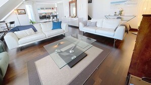 Una sala de estar moderna con dos sofás blancos, una mesa de centro de vidrio y una cocina al fondo.