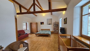 Um quarto com cama de madeira, sofá vermelho, cadeira de vime, escrivaninha de madeira, estante de livros, televisão e janela.