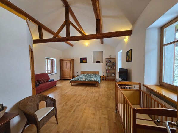 Una habitación con una cama de madera, un sofá rojo, una silla de mimbre, un escritorio de madera, una estantería, un televisor y una ventana.