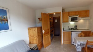 Een compacte kamer met kitchenette, een bank met kussen, houten kastjes en een deur naar een andere kamer.