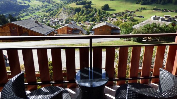 Een balkon met rieten stoelen en een glazen tafel, met uitzicht op een berglandschap met een dorp beneden.