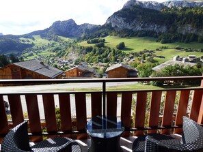Un balcón con sillas de mimbre y una mesa de vidrio con vistas a un paisaje montañoso y un pueblo en la falda de la montaña.