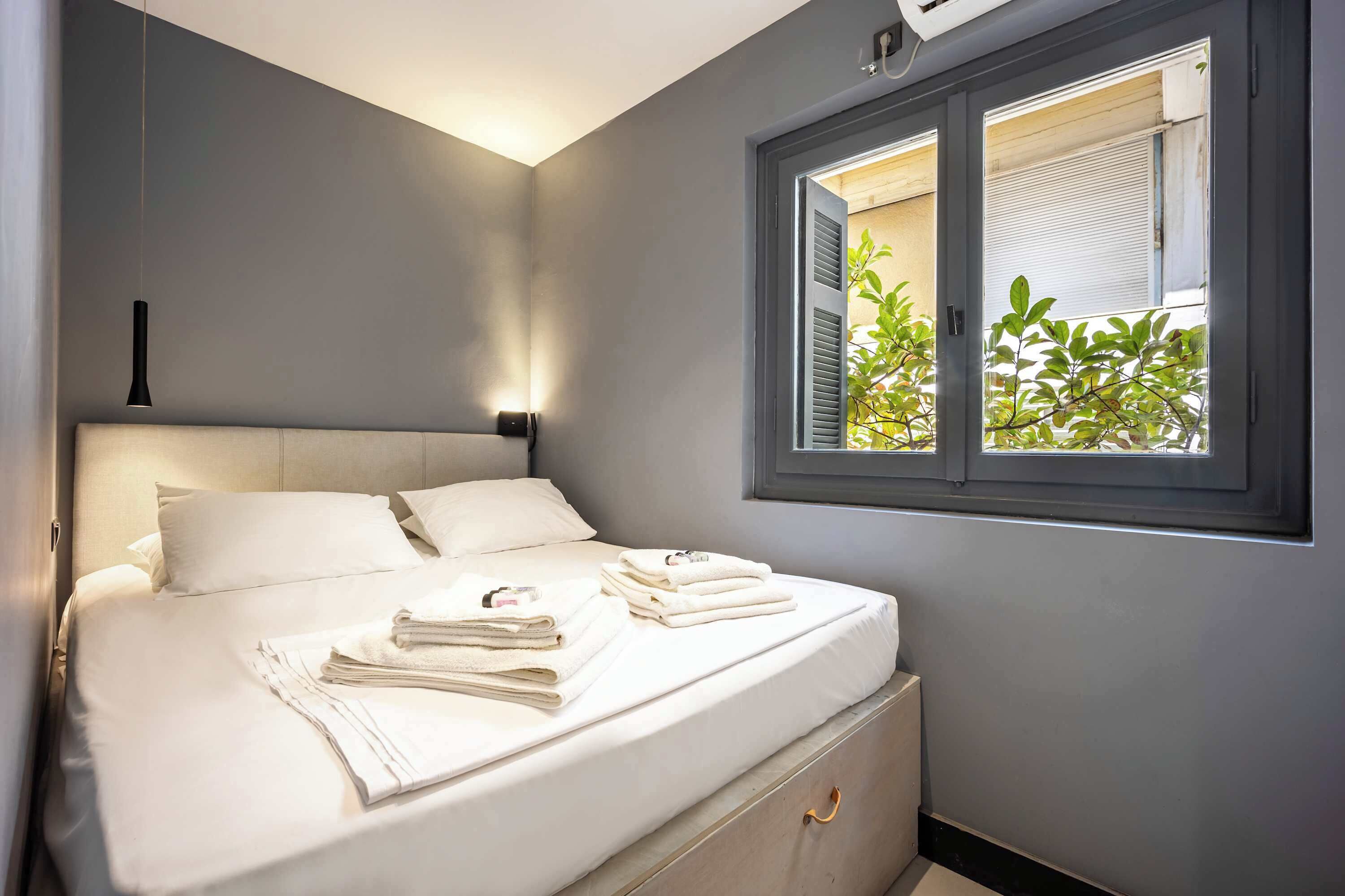 A neatly made bed with white linens and towels, a bedside table with a lamp, and a window with blinds.