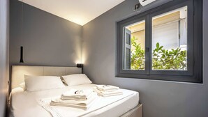A neatly made bed with white linens and towels, a bedside table with a lamp, and a window with blinds. - Well Designed 1BR in Prime Kolonaki (Athina)