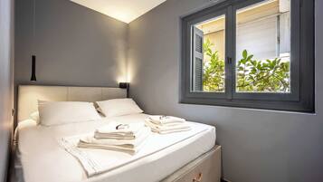 A neatly made bed with white linens and towels, a bedside table with a lamp, and a window with blinds.