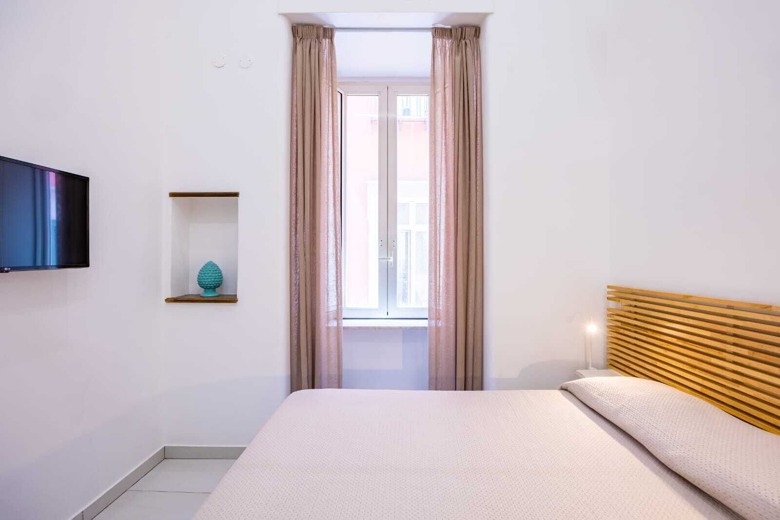 A bedroom with a bed, a TV mounted on the wall, a window with curtains, and a small shelf with a decorative item.