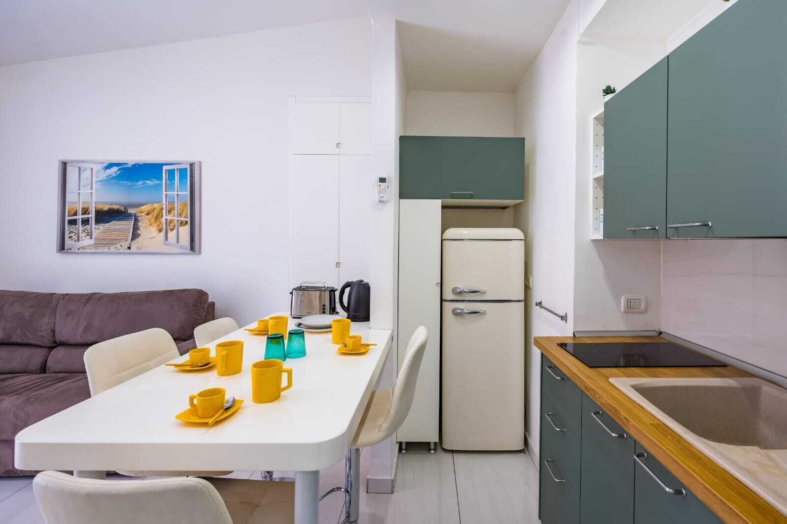 A compact living space with a dining table set with yellow cups, a kitchenette with a fridge, and a sofa.