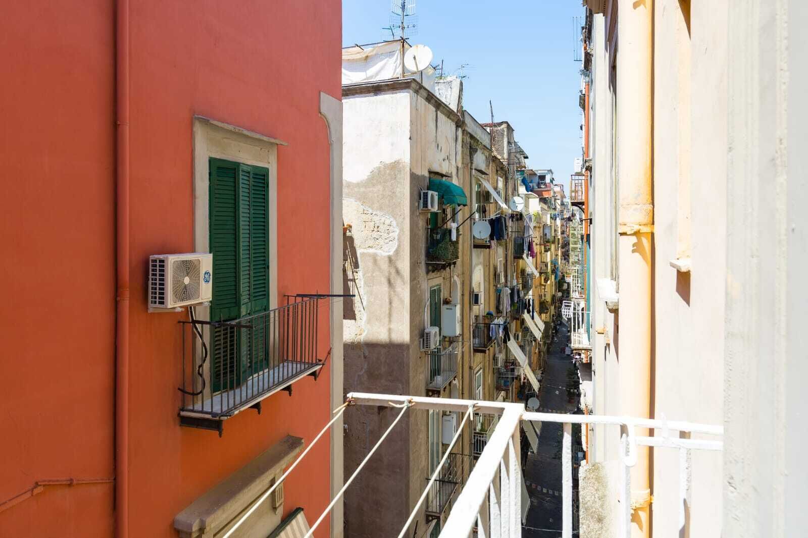 A narrow street with closely packed buildings, balconies with clothes hanging, and air conditioning units.