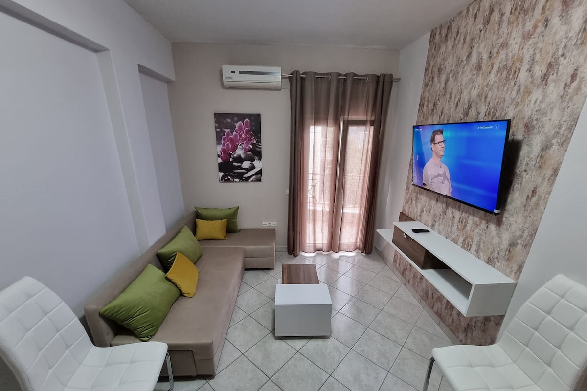 Een moderne woonkamer met een beige hoekbank, twee witte fauteuils, een salontafel en een flatscreen-televisie bevestigd aan een gestructureerde muur.