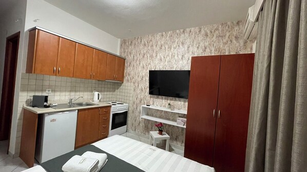 Habitación de hotel compacta con kitchenette, televisor de pantalla plana, cama con cubrecama a rayas y una mesita de noche pequeña.