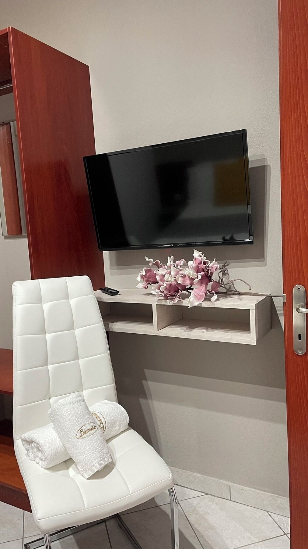 A modern bathroom with a white cushioned chair, a vanity with a mirror, a television mounted above a shelf, and a floral arrangement.
